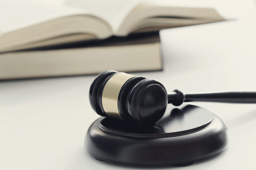 2024 Midyear Employment Law Compliance Trends 1 A wooden gavel rests on a sound block, symbolizing law and justice. In the background, an open book hints at 2024 compliance trends in employment law, offering a glimpse into the legal landscape. The image maintains a soft, neutral tone.