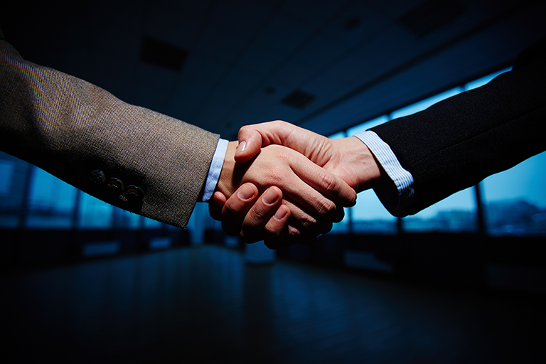 Two people in business attire are shaking hands in a dimly lit office space with large windows and a city view, symbolizing agreement at the TechServe Alliance Executive Summit, where Ceipal stands out as a proud Gold Sponsor.