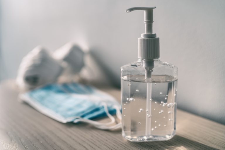 A clear bottle of hand sanitizer with a pump sits on a wooden surface, with a blue surgical mask and two white masks blurred in the background—reminders of safety measures alongside COVID-19 vaccine efforts.
